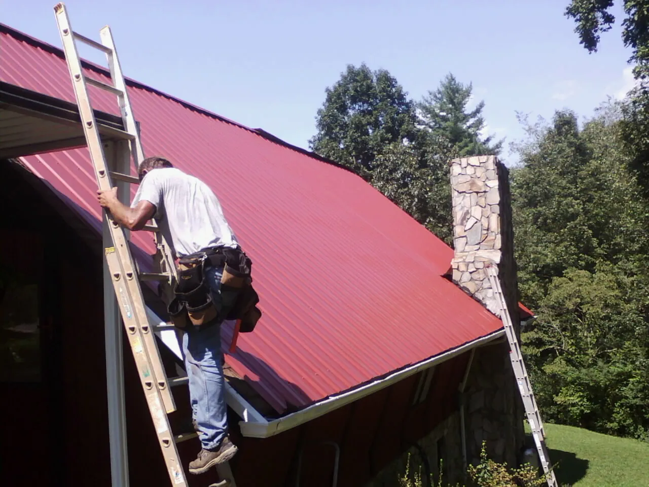 Roofer on a ladder beside a red metal roof and stone chimney — Soffit & Fascia Repair by Roofing Brothers in Bella Vista